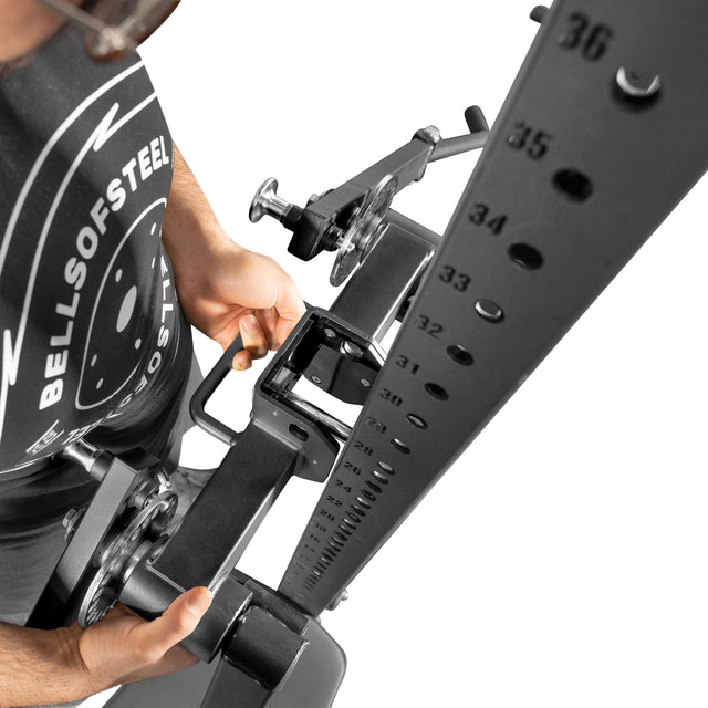 A person in a Bells of Steel shirt adjusts the Shoulder Boulder/Chest Fly Attachment, aligning numbered holes. The overhead shot highlights the adjustment mechanism of this space-saving gym equipment.