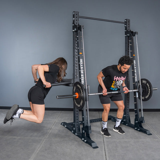 A woman does a bodyweight dip with resistance bands on the Bells of Steel USA Smith Machine - Hydra (3" x 3", ⅝" Holes), while a man prepares for a bent-over row with a loaded barbell; both wear athletic clothing.
