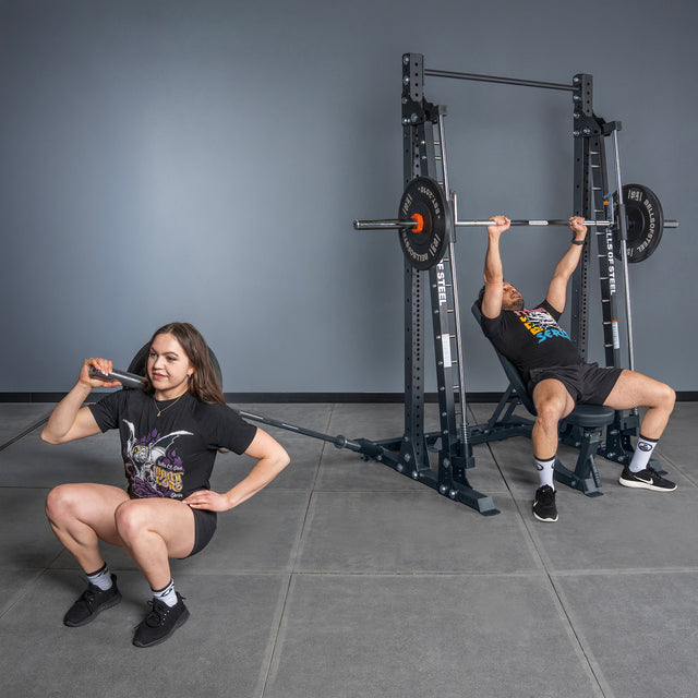 A woman squats with the Bells of Steel USA Smith Machine - Hydra (3" x 3", ⅝" Holes), while a man bench presses nearby; both are working out in a gym with gray flooring and walls.
