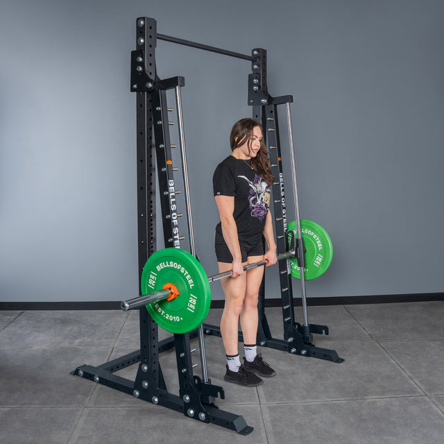 In a gym, a woman in a black T-shirt, shorts, and sneakers stands ready to lift a barbell with green plates at the Bells of Steel USA Smith Machine - Hydra (3" x 3", ⅝" Holes).