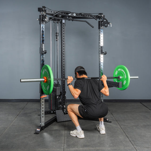 Wearing black and white shoes, a person performs a squat with green weight plates on the Bells of Steel All-in-One Trainer power rack in a home gym with gray tile flooring and a plain gray wall.