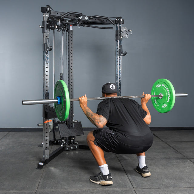 Wearing athletic gear, an individual performs a barbell squat with green weight plates inside a Bells of Steel All-in-One Trainer, set in a home gym with gray walls and floor.