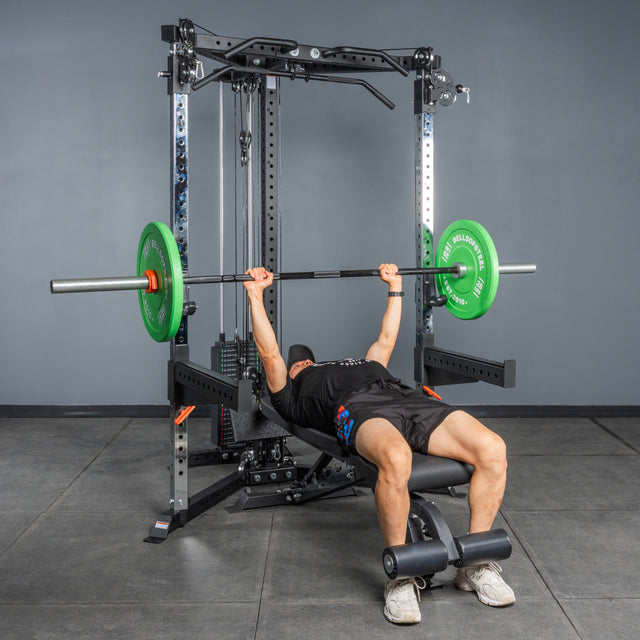 A person, dressed in a black cap, shirt, and shorts, performs a bench press with a barbell and green weights on the Bells of Steel All-in-One Trainer in a home gym.