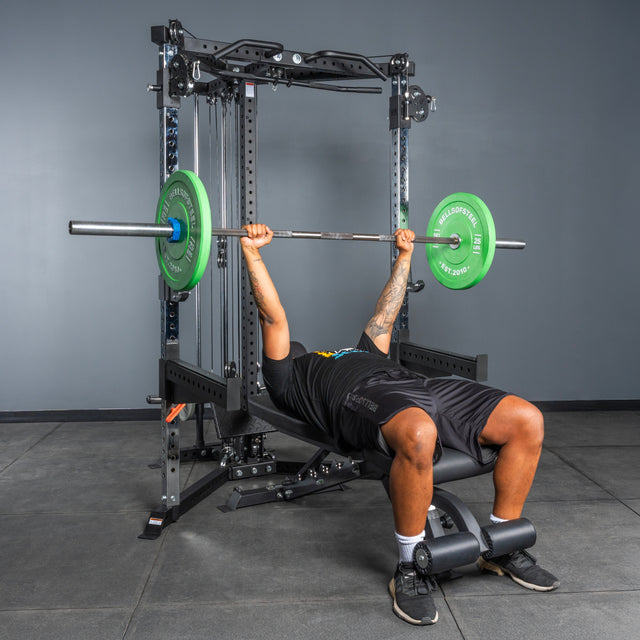 A person is bench pressing a barbell with green plates using the Bells of Steel All-in-One Trainer power rack in a gym with gray walls and black flooring.
