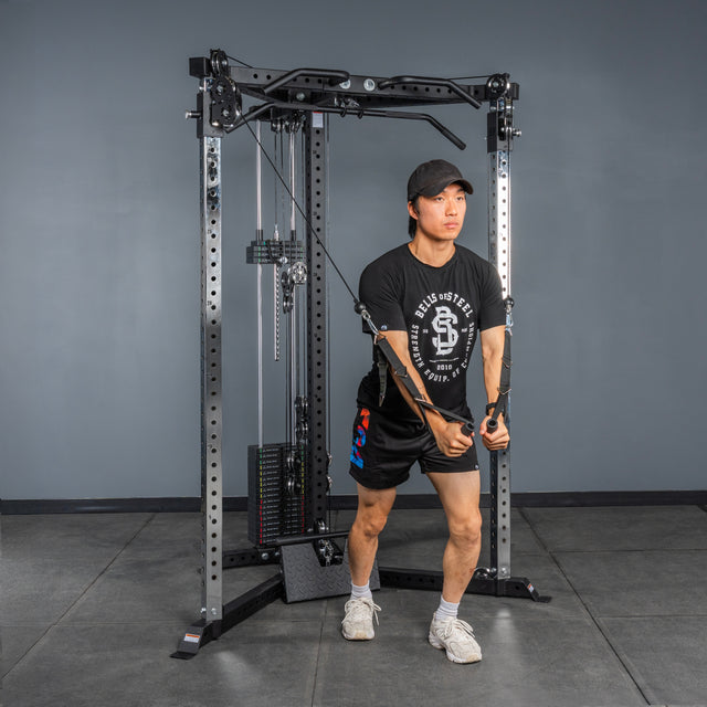 Wearing a black T-shirt, shorts, and a cap, someone performs a standing cable crossover on the Bells of Steel All-in-One Trainer in the gym, gripping both handles with arms extended forward.