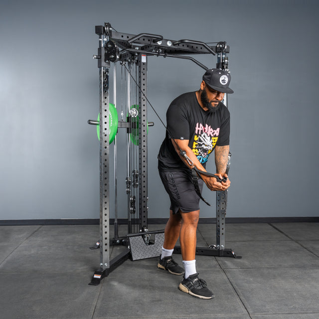 A man in a black hat, t-shirt, and shorts uses the Bells of Steel All-in-One Trainer to perform a cable crossover in a gym with gray walls and rubber flooring.