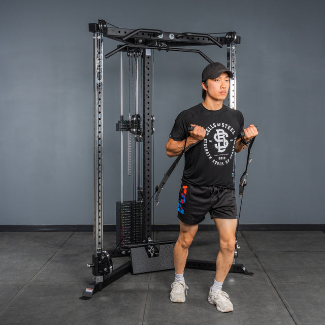 A person in black workout attire uses the Bells of Steel All-in-One Trainer, pulling handles upward in a gym with gray walls and black flooring, demonstrating the machine’s versatility.