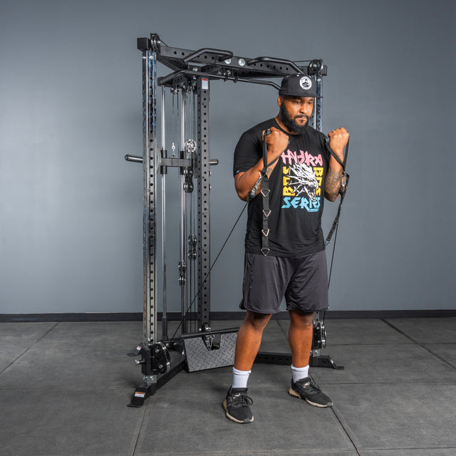 A man in athletic wear uses Bells of Steel's All-in-One Trainer with resistance bands in a gym. Standing on a black mat, he grips the bands with both hands and bends his arms to perform a strength exercise.