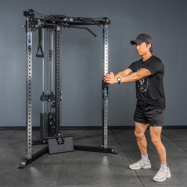 A person in black athletic wear stands with arms extended, gripping a cable handle attached to the Bells of Steel All-in-One Trainer in a home gym.