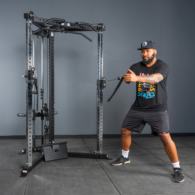 A man in athletic wear and a cap uses the Bells of Steel All-in-One Trainer at the gym, pulling a handle with both hands while standing in a slight squat on a rubber floor.