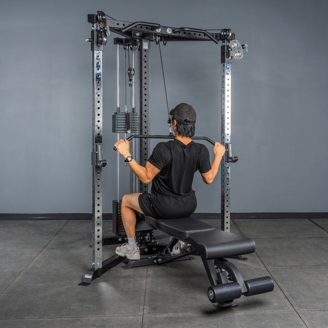 A person in black workout clothes and a cap performs a lat pulldown while seated on a bench, using the Bells of Steel All-in-One Trainer in a gym.