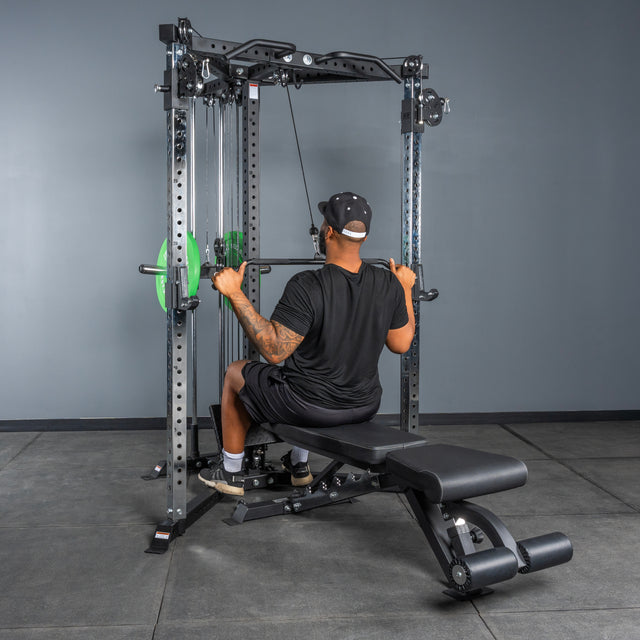 A person wearing a black shirt, shorts, and cap performs a lat pulldown on the Bells of Steel All-in-One Trainer in a gym, seated on a bench and pulling the bar down behind their neck.