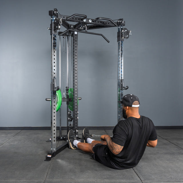 A person in black workout clothes and a cap sits on the gym floor, holding a cable row handle attached to the Bells of Steel All-in-One Trainer, getting ready for a seated cable row exercise.
