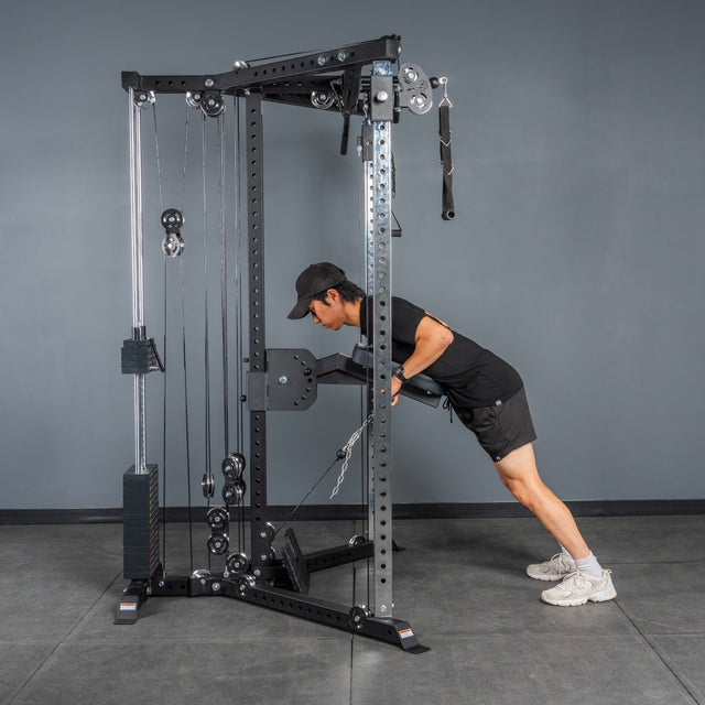 A person in a black outfit and white sneakers performs a triceps pushdown using the Bells of Steel All-in-One Trainer in a gym with gray walls and flooring.
