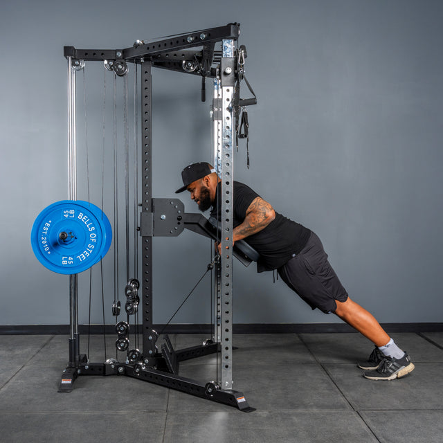 A man in athletic clothing does an incline push-up on a Bells of Steel All-in-One Trainer in a gym with gray walls and floor.