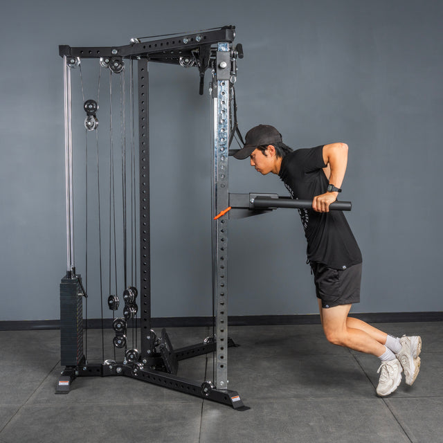 A person in athletic wear and a cap uses the Bells of Steel All-in-One Trainer to perform a tricep pushdown exercise in a gym.