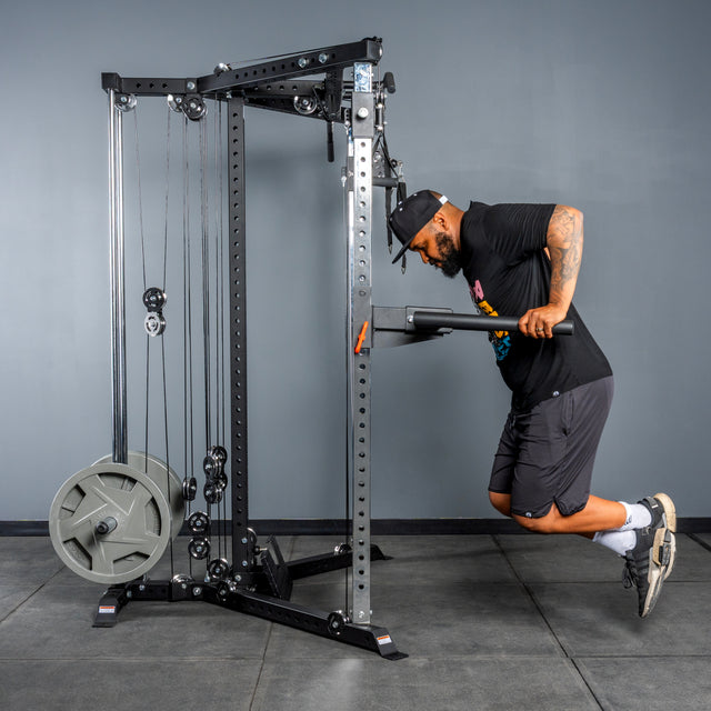 A man in athletic clothing uses the Bells of Steel All-in-One Trainer to perform assisted dips at the gym, leaning forward with bent arms and a focused expression as he grips the handles.