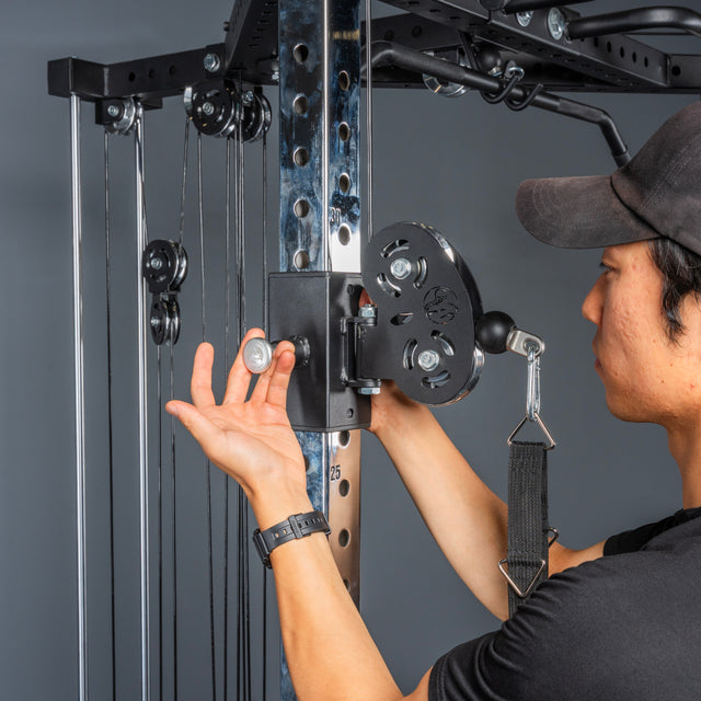 Wearing a black shirt and cap, a person uses their left hand to secure a cable pulley with a silver pin on the Bells of Steel All-in-One Trainer home gym.