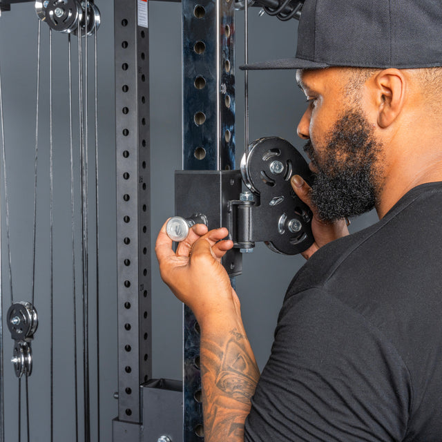 A bearded man in a black shirt and cap adjusts a metal pin on the Bells of Steel All-in-One Trainer at the gym, focusing intently on the equipment.