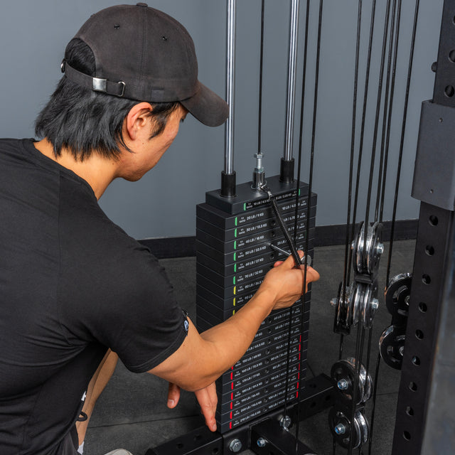A person in a black shirt and cap adjusts the weight stack on the Bells of Steel All-in-One Trainer at the gym, getting ready for a workout.