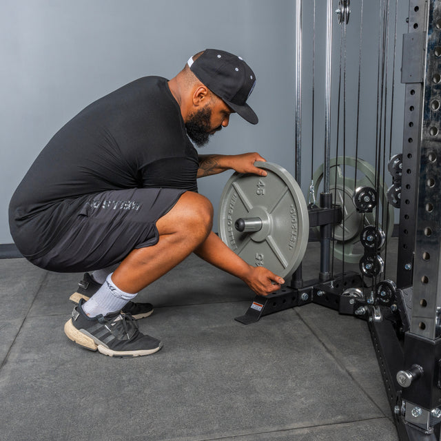 A man in athletic wear and a cap squats to load a 45-pound plate onto the Bells of Steel All-in-One Trainer in a fitness center.
