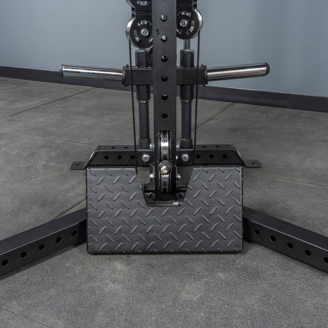 Close-up of the Bells of Steel All-in-One Trainer’s black metal base with diamond plate cover, bolts, and handles on a gray concrete floor against a plain gray wall.