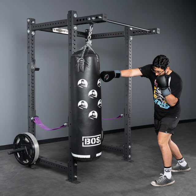 A man in black athletic wear and gray shoes punches a black BOS punching bag hanging from a Bells of Steel USA Rack Junctions - 3" x 3" setup in a home gym, with weight plates stacked on the rack.