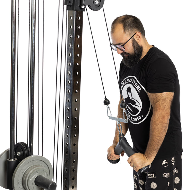 A bearded man in glasses and a black T-shirt uses the Bells of Steel Fat Bar - Tricep Pressdown on a cable machine, gripping the handle to build grip strength as weight plates are visible on the equipment.