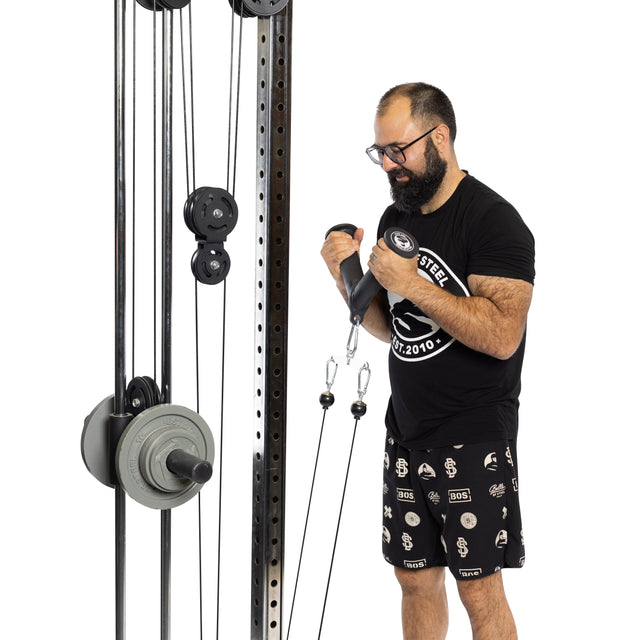 A bearded man in glasses and a black t-shirt uses the Bells of Steel Fat Bar - Tricep Pressdown on a cable machine, focusing on grip strength against a white background.