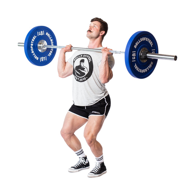 A man lifts a barbell loaded with Bells of Steel Urethane Bumper Plates to his chest, wearing a gray T-shirt, black shorts, and sneakers. He prepares for a front squat or overhead lift against a plain white background.