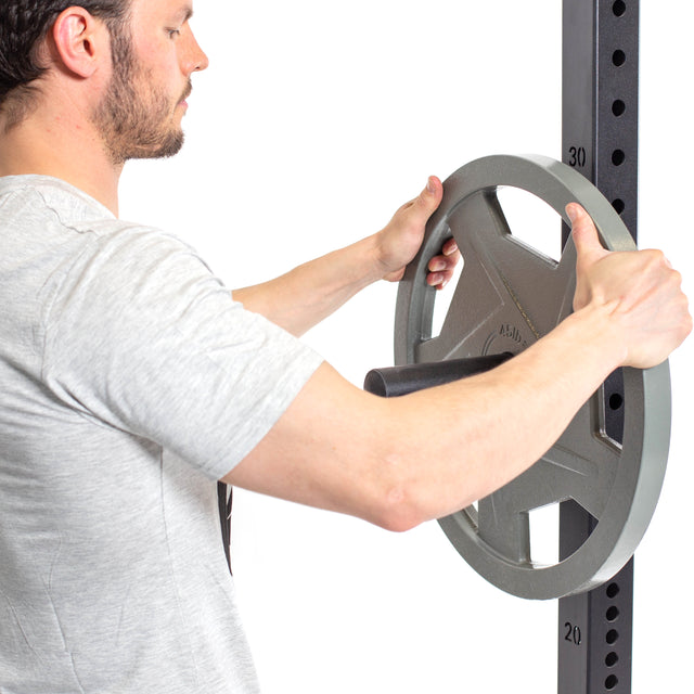 A man in a light gray t-shirt loads a large metal weight plate onto the Bells of Steel Utility Horn Rack Attachment, set against a plain white background.