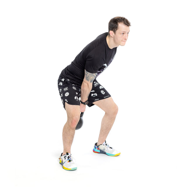 A man in a black t-shirt and patterned shorts holds a Bells of Steel 6-12KG Adjustable Kettlebell with both hands, knees and hips bent, preparing for a swing against a plain white background.