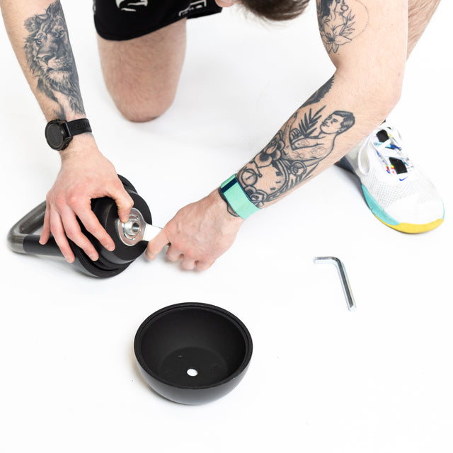 A tattooed person assembles a Bells of Steel 6-12KG Adjustable Kettlebell on a white surface, using an Allen wrench to tighten a part. A round black base and tool are nearby; only their arms and legs are visible.