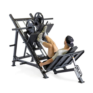 A man wearing a gray shirt, black shorts, and a black cap is using a leg press machine loaded with weight plates in a gym setting.