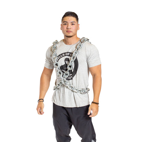 A person wearing a gray t-shirt with a graphic design and black pants stands confidently, wrapped in thick metal chains. The background is plain white.