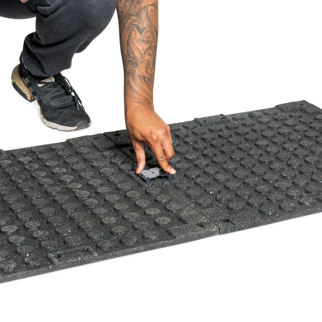 A man installs a Bells of Steel Rubber Flooring Gym Mat 24" x 24" by placing a small connector piece between tiles. Only their hand, arm, shoe, and part of the black textured mat appear against a white background.