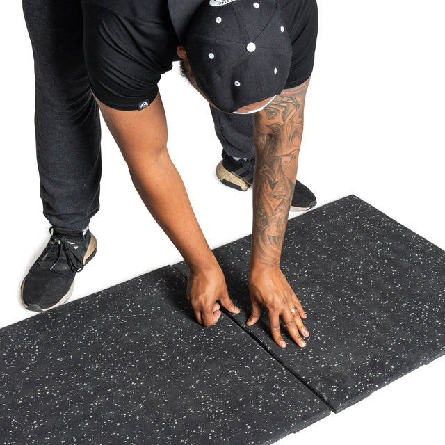 A person in black attire uses their hands to align two Bells of Steel Rubber Flooring Gym Mats (24" x 24") with a black speckled design on a white background, pressing the edges together.