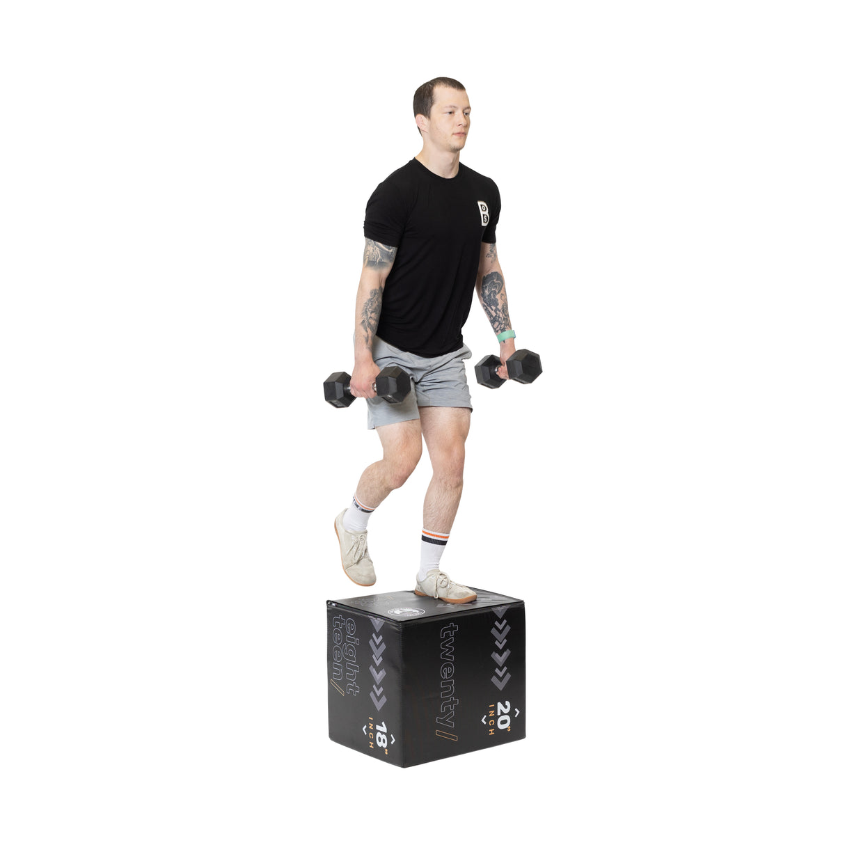 A person wearing a black t-shirt and gray shorts performs step-ups on the Bells of Steel Foam Soft Plyo Box (16 x 18 x 20), demonstrating versatile dumbbell exercises. Their tattooed arms, white socks, and beige sneakers highlight these key home gym essentials.