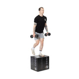 A person wearing a black t-shirt and gray shorts performs step-ups on the Bells of Steel Foam Soft Plyo Box (16 x 18 x 20), demonstrating versatile dumbbell exercises. Their tattooed arms, white socks, and beige sneakers highlight these key home gym essentials.