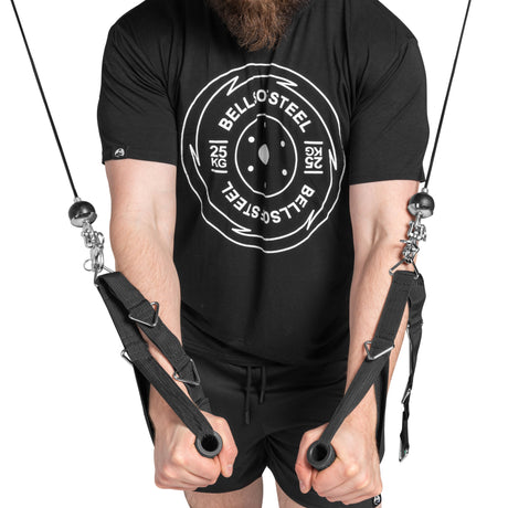 A bearded individual in a black "Bells of Steel USA" shirt and shorts uses cable machines with Swivel Shackles, gripping two handles. They're part of the vibrant home gym community.