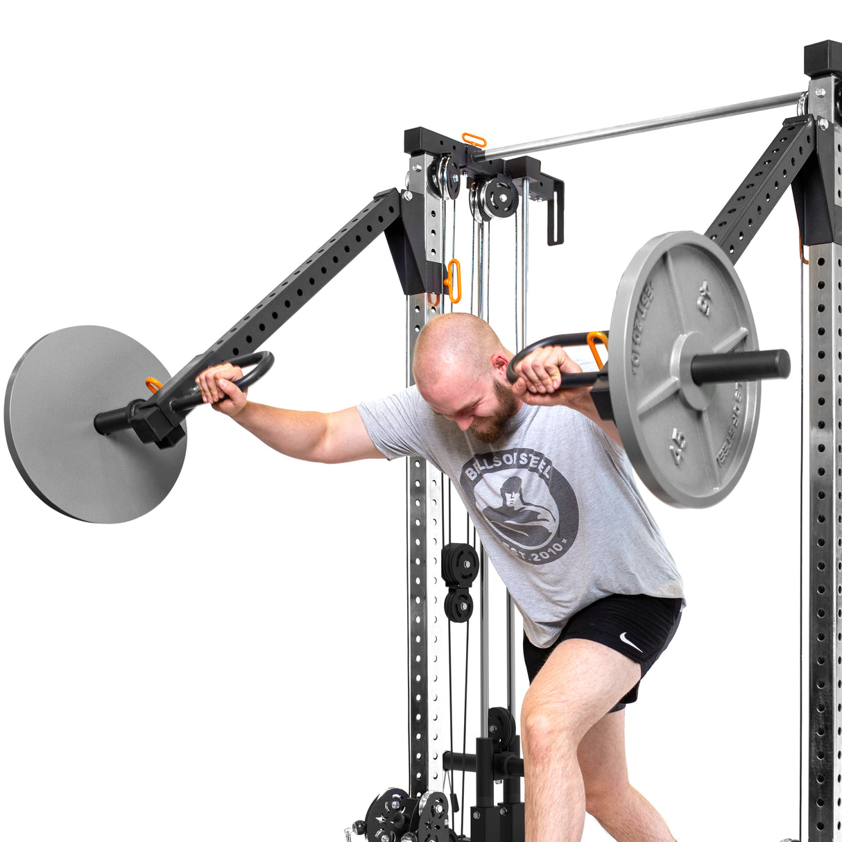 A person in a gray shirt and black shorts works out with the Bells of Steel Cable Tower Squat Stands, performing a shoulder exercise reminiscent of a home gym setup. They stand with one knee slightly bent, lifting weights against a white backdrop.