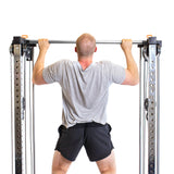 A person uses the Bells of Steel Cable Tower Squat Stands in their home gym. They wear a gray T-shirt and black shorts, facing away from the camera against a plain white background.