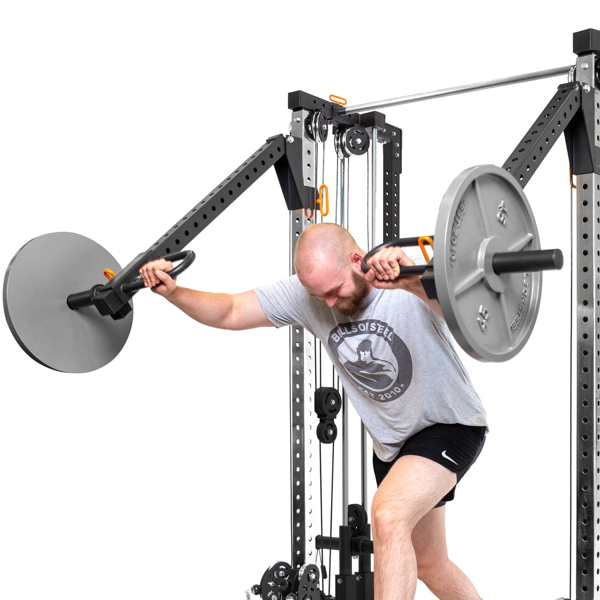 At the gym, a man in a grey T-shirt and black shorts performs a shoulder exercise using the Bells of Steel Cable Tower Squat Stands for strength training.