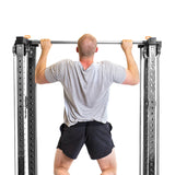 A man in gray t-shirt and black shorts performs a pull-up on Bells of Steel's Cable Tower Squat Stands against the plain white background of his home gym.