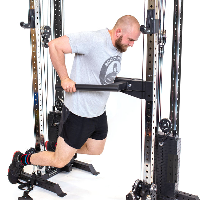 A man in a gray T-shirt and black shorts uses Bells of Steel's Cable Tower Squat Stands, showcasing its versatility in his sleek home gym. He performs dips with slightly bent legs and a forward-tilted head against a white background.