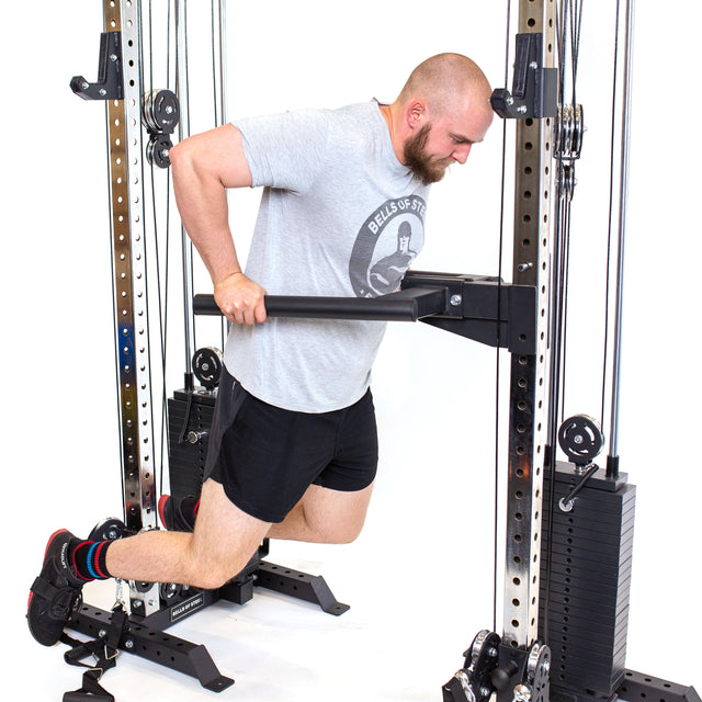 A man in gray T-shirt and black shorts exercises on a dip machine in his home gym, gripping handles with bent knees. The setup includes Bells of Steel's Cable Tower Squat Stands, a metal frame, and weight stacks that perfectly complement the cable tower nearby.