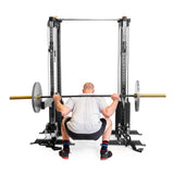 A person is squatting with a barbell on their shoulders in front of Bells of Steel Cable Tower Squat Stands. They wear a gray shirt, black shorts, and colorful socks against a plain white background, resembling the sleek aesthetic of a home gym.