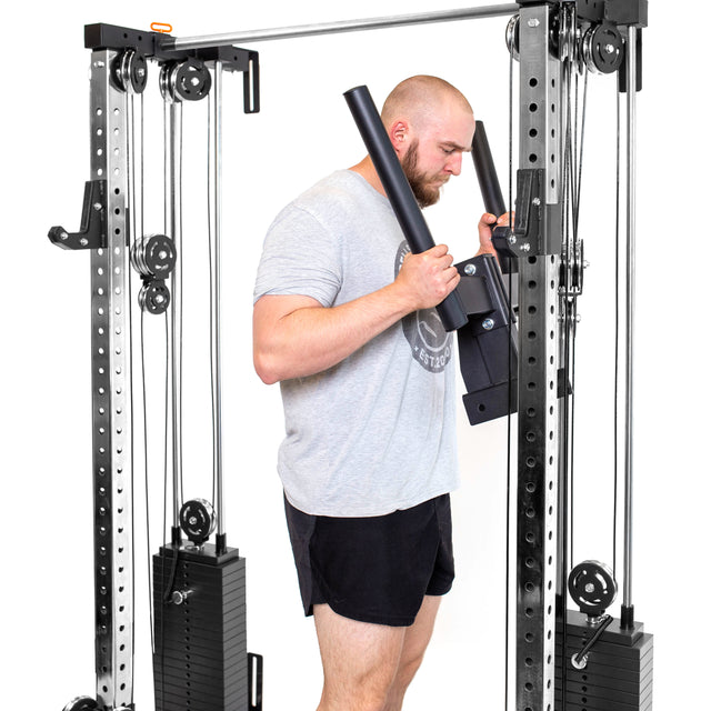 A bearded man with short hair exercises using the Bells of Steel Cable Tower Squat Stands, emulating a sleek home gym setup. In a gray T-shirt and black shorts, he stands against a white background, showcasing focus and strength with the cable-driven lat pulldown machine.