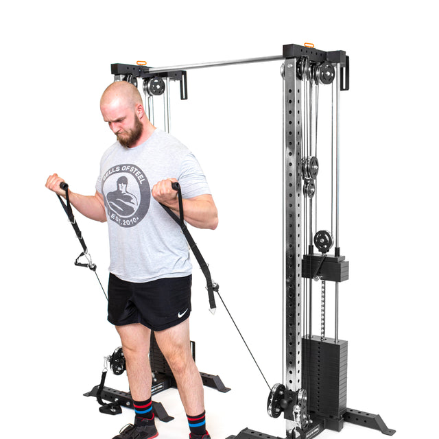 A bearded man works out with the Bells of Steel Cable Tower Squat Stands in his home gym, wearing a gray T-shirt, black shorts, and striped socks against a plain white backdrop.