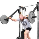 Wearing a light gray T-shirt with a distinctive logo and black shorts, a person works out on the sleek Bells of Steel Cable Tower Squat Stands, holding a barbell with weighted plates. The machine's metal frames and pulleys enhance the minimalist home gym setup.